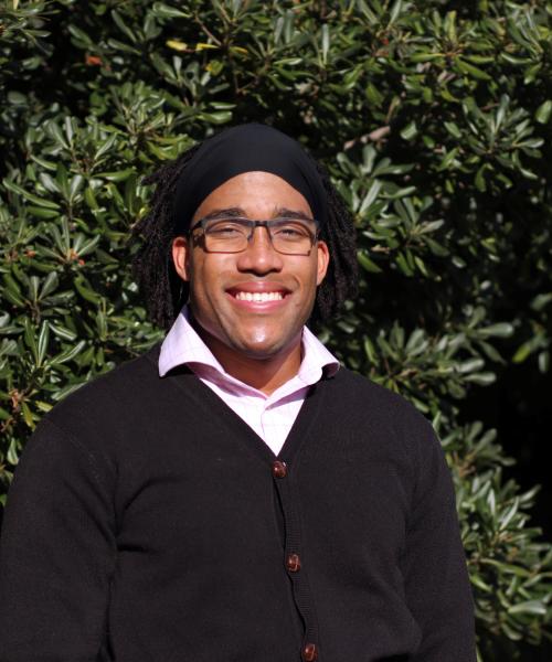 Christian in a black sweater and pink shirt standing in front of green bushes.