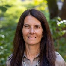 Picture of Jennifer Hoffman taken outside Miller Hall with green leafy background. Jennifer is smilling, has long brown hair and is wearing a brown blouse & light green necklace. 