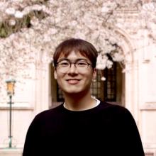 A smiling person with glasses stands in front of a building, surrounded by blooming cherry blossoms, creating a serene and joyful spring atmosphere.